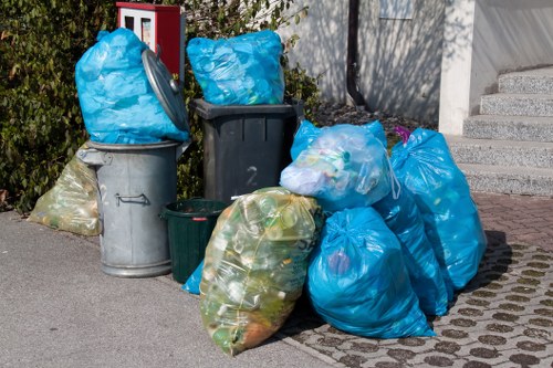 A collection of various rubbish bags and bins placed on a paved area next to a building in a residential or urban setting. The image shows multiple large plastic bags, mostly blue, filled with waste, some tied and others open to reveal their contents. There are also a few grey and green wheelie bins, some with lids closed and others open, with additional waste visible inside. The bags and bins are positioned on a mix of asphalt and paving stones, with a low garden wall, some shrubbery, and a set of concrete steps leading to an upper level of a building visible in the background. The scene appears to be a typical disposal area for rubbish collection, relevant to commercial waste removal services operating in Temple or nearby areas, emphasizing the importance of professional waste management for business premises.