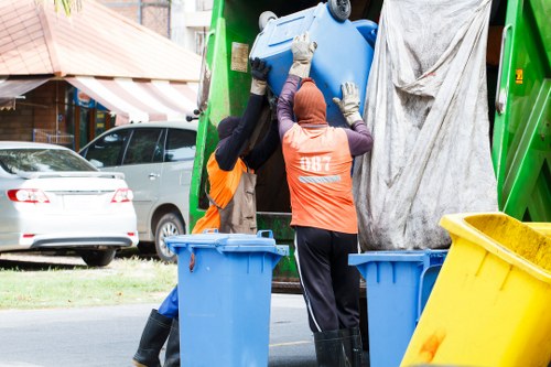 Two waste collection workers wearing high-visibility orange vests and gloves are actively lifting and emptying a large blue plastic bin into the back of a green rubbish collection vehicle parked on a residential street, with an older blue barrel container in the foreground and a yellow bin to the right. The scene takes place in a suburban area, with parked cars and houses with pitched roofs and front gardens visible in the background. The workers are positioned centrally, focused on the task, while the vehicle's rear is open, revealing its internal loading area. The environment is daytime with natural light, providing a clear view of the textured plastic bins, the metal structure of the waste truck, and the surrounding pavement. This image illustrates the process of commercial or domestic rubbish collection, typical of waste removal services offered by companies like Commercial Waste Removal Temple, serving local communities including nearby towns and postcode areas within the region.