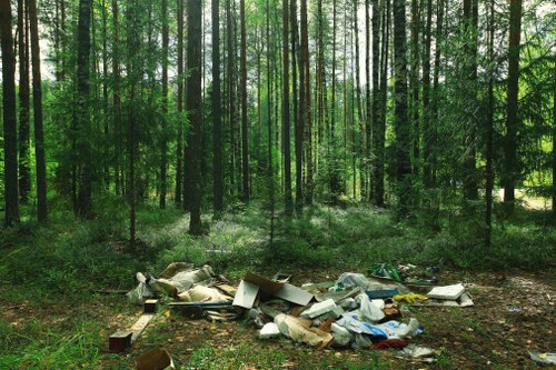 A collection of discarded rubbish scattered across a patch of ground in a wooded area, with tall, slender trees in the background. The rubbish includes various materials such as cardboard boxes, plastic bags, foam pieces, and other miscellaneous waste, some of which are partially flattened or crumpled. The waste appears to be left in an open space, with natural light filtering through the green foliage, highlighting the contrast between the natural environment and the litter. This scene exemplifies improper waste disposal, which local rubbish removal services like Commercial Waste Removal Temple may help address to maintain cleanliness and environmental health in the area around Temple and its postcode region.