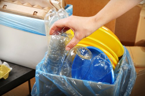 A close-up view of a person's hand holding a clear plastic water bottle filled with water, positioned above a large, open blue recycling bag lined with a transparent plastic liner. Inside the bag, there are several colored plastic containers, including a yellow lid, a blue lid, and other assorted pieces of plastic waste. The background shows a section of an indoor space with a cardboard box and a small black table. The scene depicts the sorting and disposal of recyclable plastics, highlighting waste management practices typical of commercial rubbish removal services in Temple, with an emphasis on environmentally conscious waste segregation.